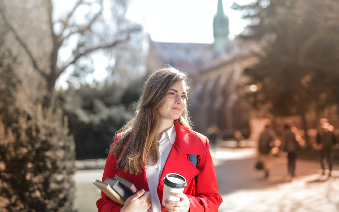 Młoda kobieta z kubkiem kawy i książką przed budynkiem uczelni.